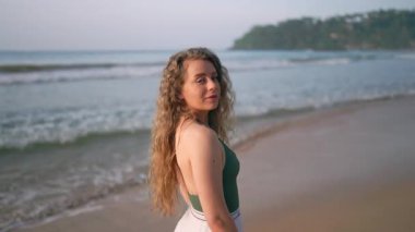 Dişi, sakin deniz kıyısından zevk alıyor, parlak saçlar sergiliyor, seyahat keyfine örnek oluyor. Yeşil mayo giymiş kıvırcık saçlı kadın tropik sahilde yürüyor, dalgalar yavaşça kıyıdan geliyor..