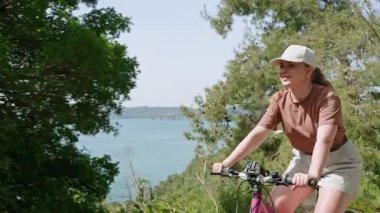 Bisikletle aktif seyahat macerası. Kadın kozalaklı ormanda bisiklet sürüyor, kıyı manzarasından zevk alıyor. Dişi bisikletçi doğayı keşfediyor, deniz manzarası görünüyor. Sağlık, çevre dostu turizm..