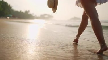 Solo gezginler sakin denizin tadını çıkarırlar, altın saat yürüyüşünün. Çıplak ayaklı kadın gün batımında kumlu sahilde elinde hasır şapkayla yürüyor. Ayaklar birbirine değecek, güneş ıslak kumu yansıtacak. Özgürlük, yaz tatili titreşimleri.