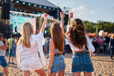 Günlük yaz kıyafetleri, canlı performans titreşimleri için sahnede toplanan enerjik kalabalık. Üç kadının kumlu sahilde müzik festivalinin keyfini çıkarırken, güneşli gökyüzünün altında içkileri yudumlarken..