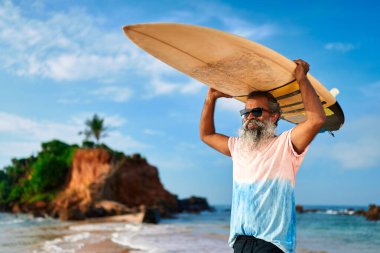 Tropik sahilde sörf tahtası olan yaşlı bir etnik adam. Güneş gözlüklü beyaz saçlı erkek sörfçü kafasının üstünde tahta tutuyor. Aktif yaşam tarzı yaşlı yetişkinler. Güneşli, mavi gökyüzünün altında okyanus dalgaları. Emeklilik maceraları