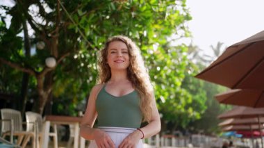 Gülümseyen bayan tropik cennette geziniyor. Parlak genç kadın deniz kenarındaki tatil beldelerinin tadını çıkarıyor. Güneşle öpülmüş model sahilde yürüyor, canlı bir tatil atmosferini emiyor. Seyahat, rahatlık ve sıcaklık..
