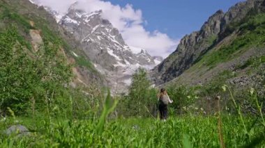 Svaneti dağlarında Chalaadi buzulu patikasında yürüyüş yapan bir kadın. Dişi yürüyüşçü, karlı tepelere doğru zengin yeşil patika boyunca yürüyor. Turist, doğa manzaralı milli parkı geziyor. Yavaşla