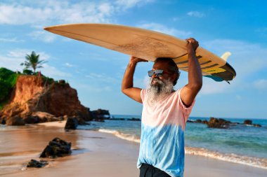 Sörf tahtası olan yaşlı adam kumlu sahilde yürüyor. Yaşlı sörfçü aktif yaşam tarzını benimsiyor. Olgun erkek, okyanus dalgalarından hoşlanır. Gümüş saçlı sporcu sörften zevk alıyor. Sağlıklı yaşlanma enerjisi.