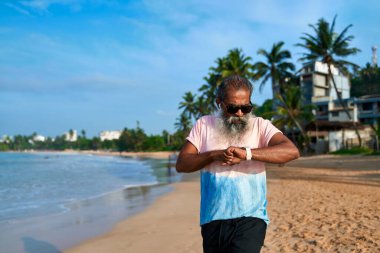 Kıdemli adam tropik plajda akıllı saat kullanır. Sakallı dedeme kulaklık, çek takip cihazı. Modern teknoloji, aktif yaşam tarzı, okyanus geçmişi. Yaşlılar dijital yenilikçiliğin keyfini çıkarıyorlar.
