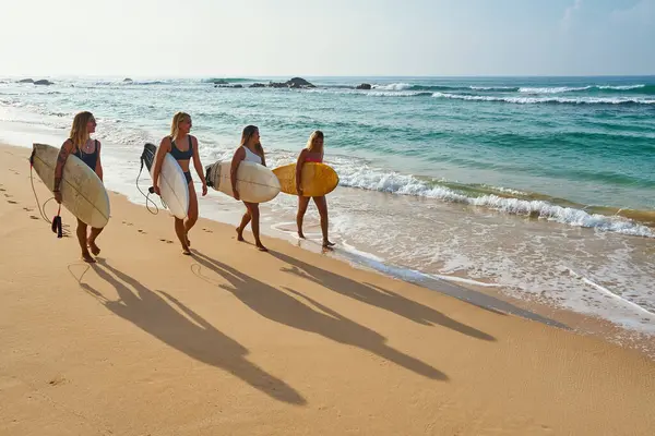 Dört kadın kumlu sahilde ellerinde sörf tahtaları ile yürüyor. Dişi sörfçüler, siluetler kumun üzerine uzun gölgeler bırakırlar. Okyanus dalgaları yakınlarda yavaşça kırılır. Açık gökyüzü ve ufuk. Deniz havası, spor aktiviteleri..