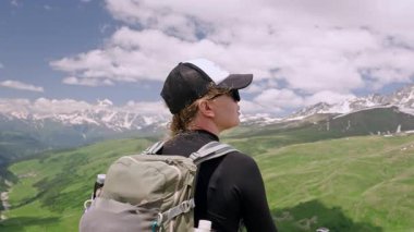 Kadın yürüyüşçü dağ manzarasından hoşlanır. Sırt çantaları, yürüyüş direkleri, şapka ve güneş gözlükleri. Yeşil tepenin üstünde duruyor. Karlı Kafkaslar arka planda zirve yapar. Seyahat, yaz yürüyüşü Svaneti bölgesinde macera. Slomo