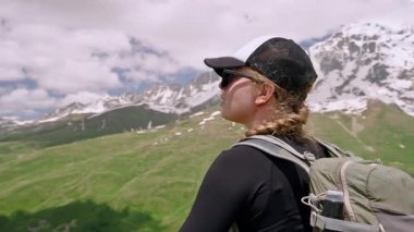 Kadın yürüyüşçü, Büyük Kafkasya 'daki Svaneti dağlarının manzarasına hayran. Sırt çantalı bir kadın doğa yürüyüşü yapıyor. Dağ yürüyüşü macerası. Doğa yolculuğu tecrübesi. Yeşil tepeler karla kaplı zirveler. Yavaşla