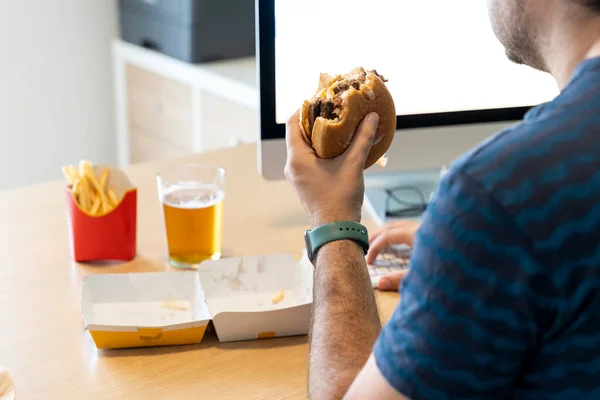 Man eating a hamburger, fries and drinking a beer while working in his office in front of the computer