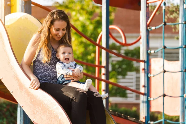 Anne bebeğiyle birlikte güneş batarken parkta kayıyor. Arka planda bazı konut binaları