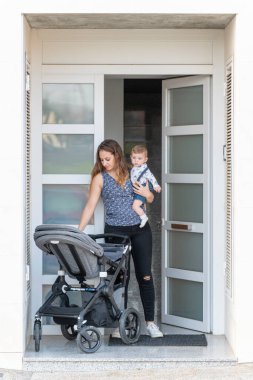 Genç bir anne ve oğlunun dikey portresi. Evlerinin kapısında. Bebek arabasıyla gitmeye hazır.