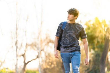 Kıvırcık saçlı genç esmer adam arka planda ağaçlarla parkta yürüyor.