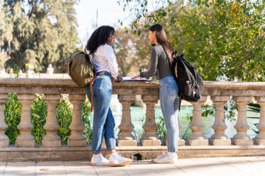 Sokakta, arka planda ağaçlar olan defterlerine bakan iki genç kız öğrenci.