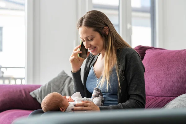 stock image Portrait of a young mother on the sofa in the living room talking on the phone with a baby in tow