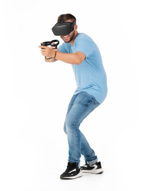 Genç bir adam beyaz bir arka planda VR gözlükleriyle oynuyor sanki bir silahla ateş ediyormuş gibi.