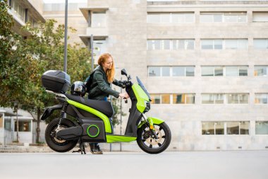 Kızıl saçlı kadın yeşil elektrikli motosikletinin anahtarlarını sokaktan içeri sokuyor.