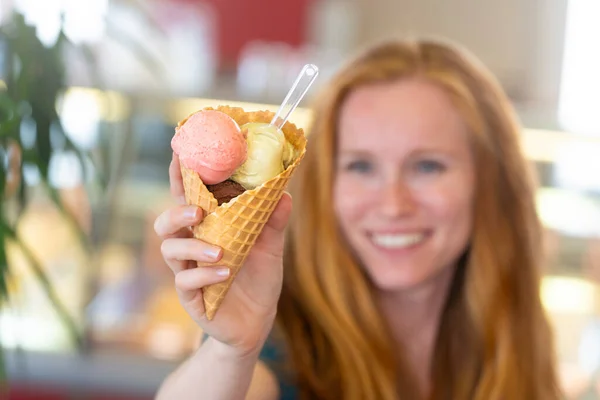 Redhead woman showing an ice cream with three flavors to the camera in an ice cream shop