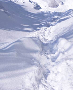 Güneşten parlayan kar beyazı kar yığınının üzerindeki insan ayak izleri, ani bir kar yağışı, karın zıt bir arkaplanı ve öğle vakti gölgeli ayak izleri; dikey kar dokusu;