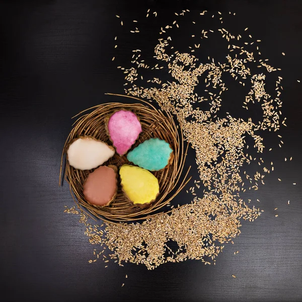Multi-colored cakes (turquoise, beige, pink, yellow and caramel) served on a wicker dish around scattered grains on a black background, square, top view, flat lay