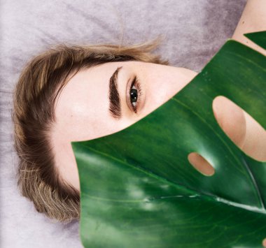 Genç bir kadının yüzü, güzellik salonunda yarım bir canavar yaprağıyla kaplıdır. Derin temizlik, spa rahatlama, kare, üst manzara, yakın çekim.
