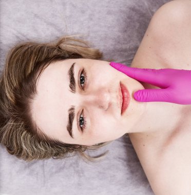 Güzellik salonunda piercingli bir kız elini pembe eldivenlerle uzatıp yüzüne dokunuyor. Cilt hastalığının teşhisi, yüz derisi ile ilgili bakım ve prosedürler. Kare, üst manzara, yakın çekim