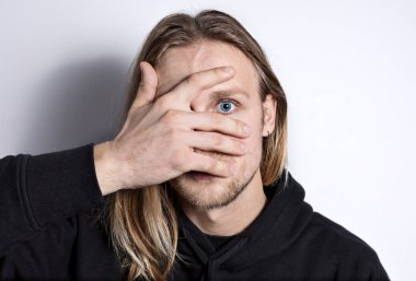 Sade bir arka planda mavi gözlü, uzun saçlı, gerçek sarışın bir adamın portresi, parmaklarından kameraya bakmaktan korkan, yüzü eliyle kaplıdır;