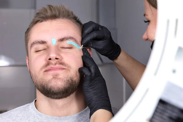 Depilation of the face with warm wax. A female eyebrow stylist applies melted wax to the eyebrows of a young unshaven blond man. Cosmetology for men, professional skin and hair care.