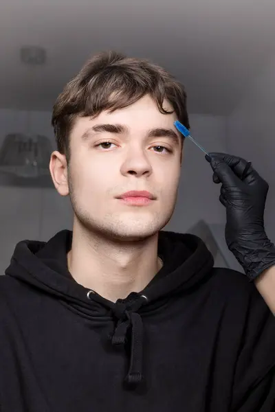 Real handsome young man with brown eyes during eyebrow correction. Eyebrow stylist models and shapes eyebrows with a blue brush.Cosmetology for men, professional hair and skin care, vertical, close-up