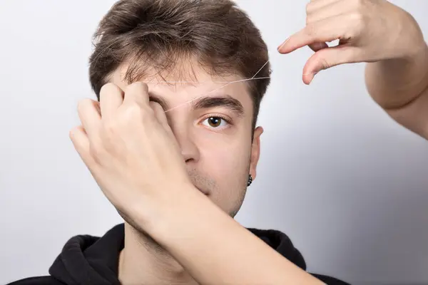 Real young man with brown eyes during eyebrow correction.The eyebrow stylist shapes the client\'s eyebrows with a thread.Cosmetology for men,professional skin and hair care, threading procedure.Closeup