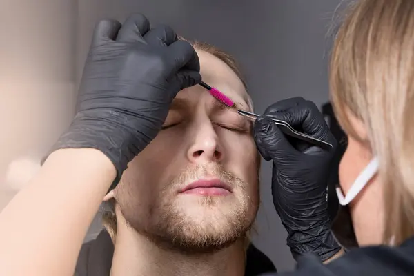 Real young man with closed eyes during eyebrow correction. The eyebrow stylist shapes the eyebrows with tweezers and a brush. Cosmetology for men, professional skin and hair care, close-up
