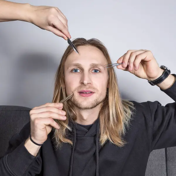Real smiling young blonde with blue eyes during eyebrow correction. Three hands point eyebrow tweezers towards his face. Cosmetology for men, professional skin and hair care, square, close-up