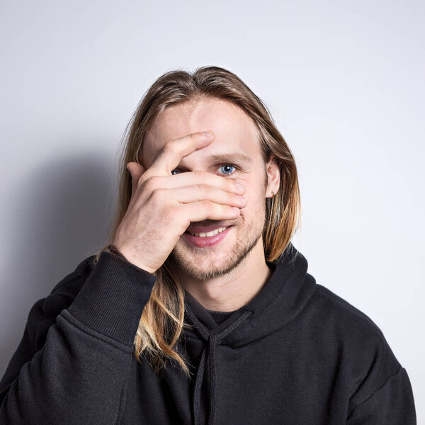 Portrait of a real smiling young blond man with blue eyes and long hair on a plain background, looking through his fingers straight into the camera, his face is covered with his hand; square