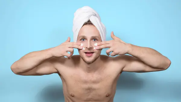 An expressive emotional topless young man with a white towel on his head smears white strips of cream over his face with his fingers.Cosmetology and home skin care for men.Close-up, blue background.
