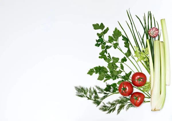 A green leafy vegetables (parsley, dill, onion, celery stalks) with tomato and garlic on a white background, isolate.Healthy food concept, fragrant herbs, organic food, flat lay, copyspace on the left