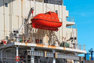 Sağlam bir kancayla büyük bir gemiye güvenli bir şekilde bağlı kırmızı bir filika, hareketli bir limanda beyaz gemiyle ve açık mavi gökyüzüyle çelişiyor, güvenli ve maceralı bir yolculuk vaat ediyor..