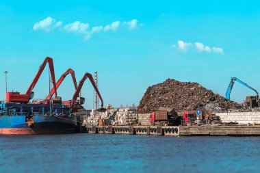 Kalabalık bir hurdalığa büyük bir kargo gemisi yanaştı. Büyük bir vinç hurda metali büyük bir yığına boşaltıyor. Bölgede çeşitli sanayi operasyonları sürüyor.