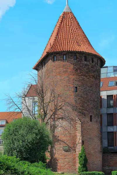 The centuries-old red brick tower with a cone-shaped red tile roof stands in a city setting. It has small windows evenly spaced along the sides and is surrounded by trees.