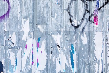 Bu resim yıpranmış bir sokak posterini gösteriyor. Kağıt ve grafiti katmanları soyulmuş. Poster, renkli ve dokulu bir arkaplan oluşturan reklamların kalıntılarıyla kaplı..