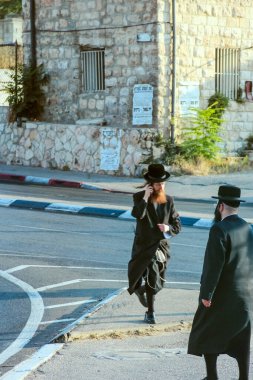 Jerusalem, Israel - July 13, 2015.In the vibrant streets of Jerusalem, two Orthodox Jewish men wearing traditional black hats and coats, one engaged in a phone conversation while the other looks on clipart