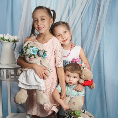 Üç mutlu çocuk, iki kız ve bir oğlan, doldurulmuş hayvanlarla birlikte parlak mavi bir perdenin önünde duruyorlar. Gülümsemelerinde saf neşe ve masumiyet göstererek kameraya mutlu bir şekilde ışıldıyorlar..