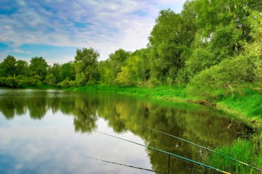 Bir nehrin kıyısına iki olta atılır. Su sakin ve durgun, gökyüzünü ve kıyıdaki ağaçları yansıtıyor. Gökyüzü bulutlu ve hafif bir rüzgar var..