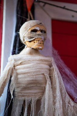 Bu yırtık pırtık beyaz bir kumaşa sarılmış bir iskeletin ürkütücü Cadılar Bayramı süslemesi. Kırmızı bir kapıda asılı ve çok ürkütücü görünüyor. Perili ev ya da Cadılar Bayramı partisi için mükemmel..