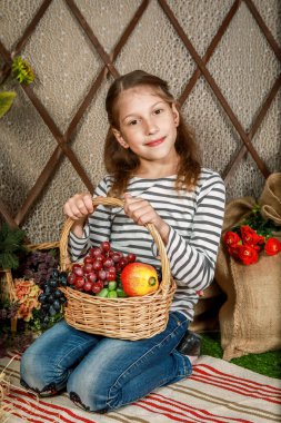 Genç bir kız, çeşitli taze meyve ve sebzelerle dolu bir sepetin yanında diz çöker ve yıpranmış ahşap bir çitin arkasından seyirciye bakarken neşe saçar..
