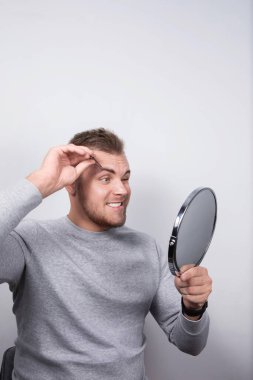 20 'li yaşlarda beyaz bir adam aynaya bakarken cımbızla kaş yoluyor. Şaşırmış görünüyor ve sol elinde gri bir kazak ve siyah bir saat var..