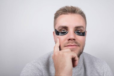 Siyah göz bantları, sarı saçlar ve kısa sakallı genç bir adam yoğun bir şekilde kameraya bakıyor, düşünceli bir şekilde. Gri gömleği biraz özgünlük katıyor..