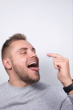 Gri tişört giyen sakallı bir adam parmağında diş macunuyla ağzını gösterdiğinde şaşırmış görünüyor. Sol elinde bir saat var ve açık gri bir arkaplanda duruyor..