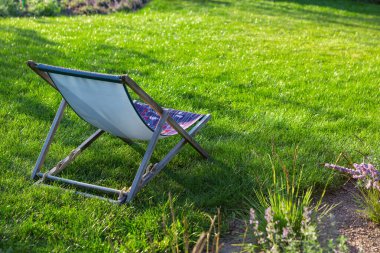 Güverte sandalyesi, sakin bir bahçe ortamındaki yemyeşil çimenlere dayanır. Parlak çiçekler ve yumuşak güneş ışığı huzurlu atmosferi güçlendirir, doğanın rahatlamasını ve eğlenmesini sağlar..