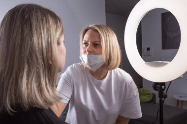 Bir makyaj sanatçısı bir kadının yüzünü ustalıkla inceler. Zarif bir beyaz önlük giyen kadın, dönüşümün iyi aydınlatılmış bir güzellik salonunda gerçekleşmesinden memnun görünüyor..