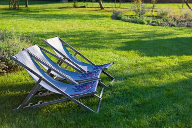 Üç rahat şezlong, yemyeşil bir çimenliğin üzerinde, yemyeşil bahçelerle çevrili. Güneş ışığı rahatlatıcı bir ortam yaratır, doğada dinlenmeye ve rahatlamaya davetiye çıkarır..