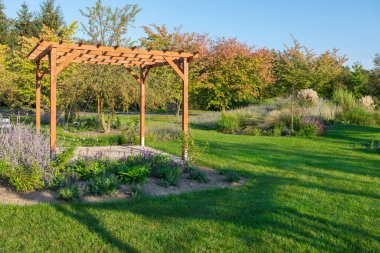 Sakin bir bahçe, merkezinde çeşitli bitki ve ağaçlarla çevrili ahşap bir pergola barındırır. Parlak güneş ışığı yemyeşil yeşilliği vurgular, barışçıl bir atmosfer yaratır..